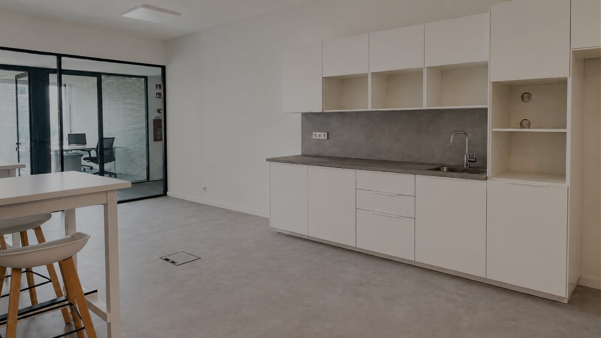 Copa equipada da MaiSpace com armários brancos, bancada, lava-louça, zona de refeições e divisórias em vidro que separam o espaço das áreas de trabalho.